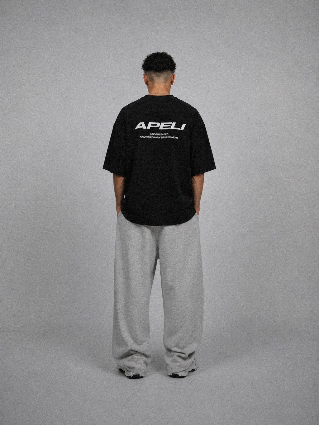 Person wearing a black t-shirt with 'APELI' logo on the back, standing against a gray background