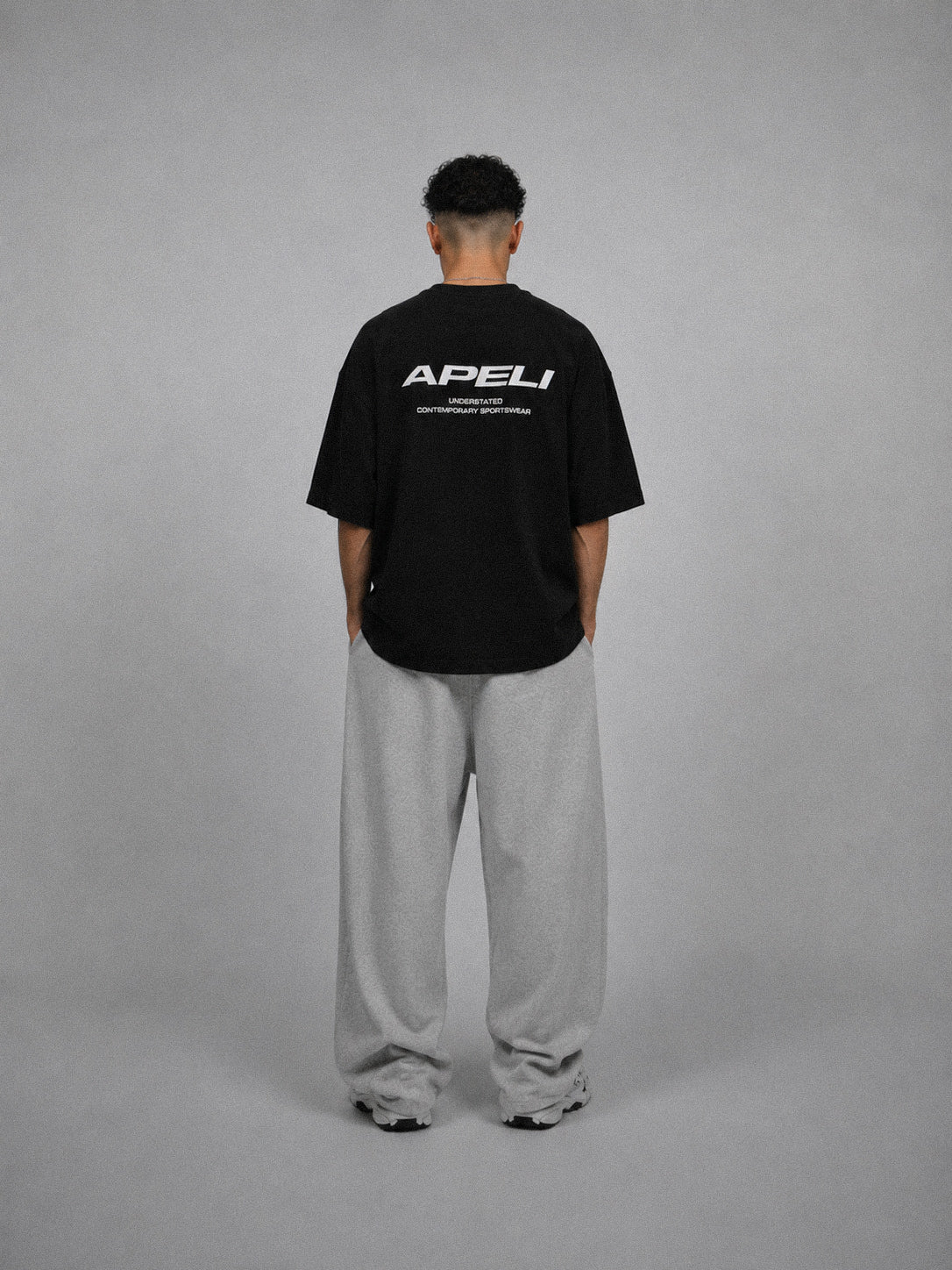 Person wearing a black t-shirt with 'APELI' logo on the back, standing against a gray background